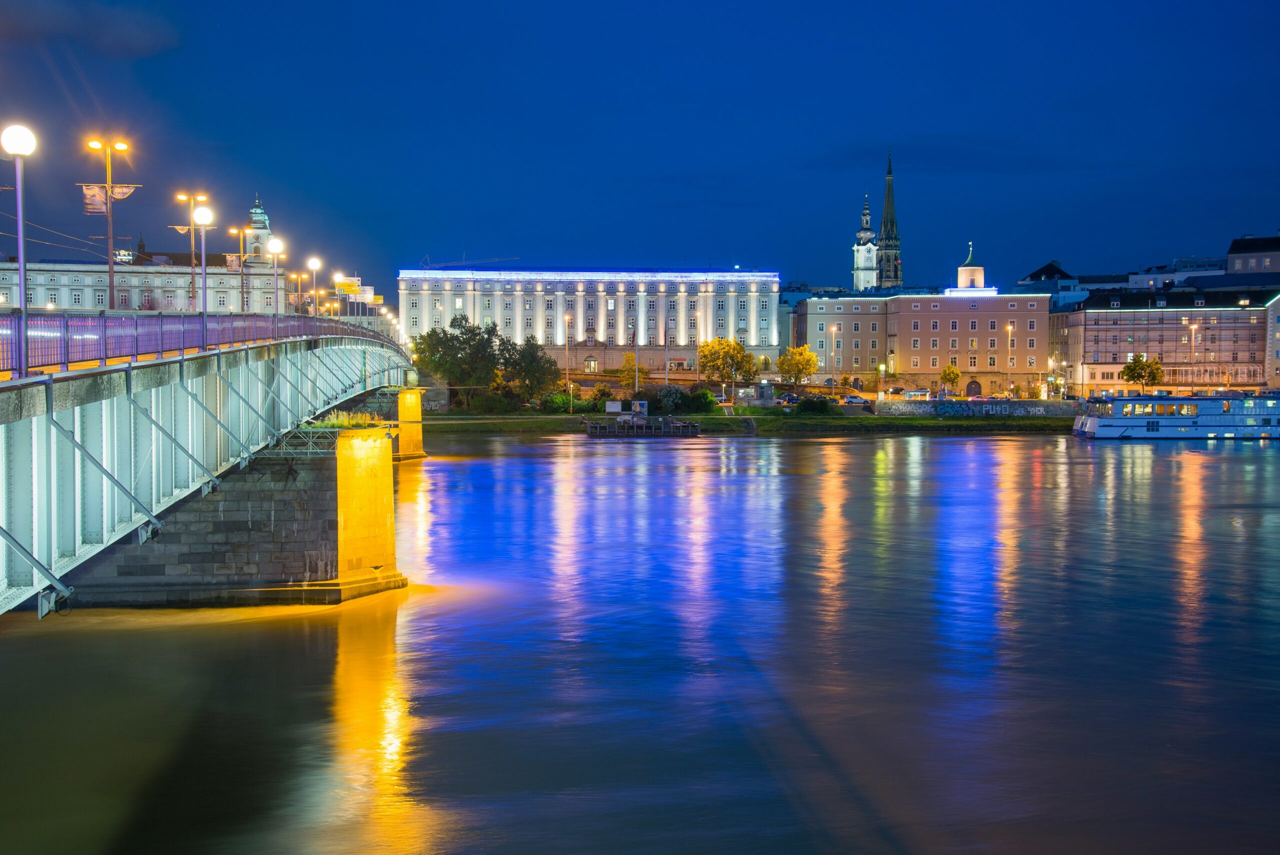alain-bonnardeaux-S_KH3f2JB2k-unsplash Blick auf das nächtlich beleuchtete Linz mit der Nibelungenbrücke und dem Donauufer, im Hintergrund das markant angestrahlte Brucknerhaus und die Altstadt.